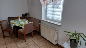 Een kamer met een eettafel, stoelen, een radiator, een raam met gordijnen en een potplant.