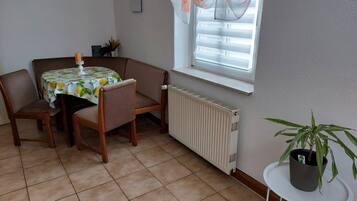 Ein Zimmer mit Esstisch, Stühlen, Heizkörper, einem Fenster mit Vorhängen und einer Topfpflanze.