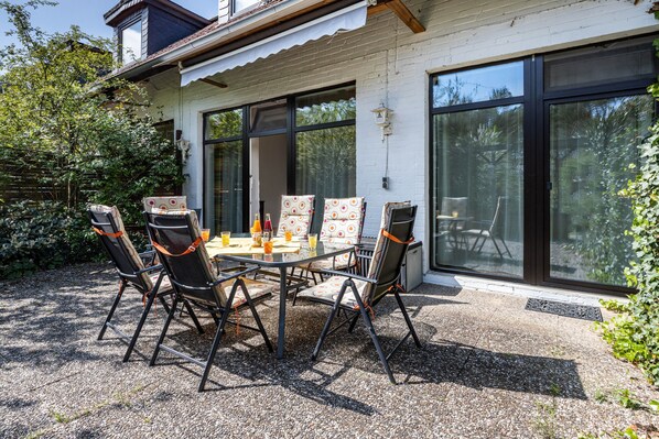 A patio with a table and chairs, a white building with large windows, and a view of trees.