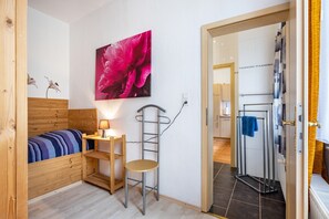 A room with a bed, a desk with a lamp, a chair, a ladder, a large flower painting, and a bathroom visible through an open door.
