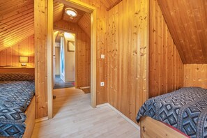 Interior de madeira com teto inclinado, duas camas com roupa de cama estampada e um corredor que leva a outro quarto.