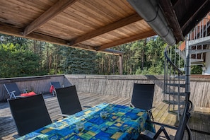 Um deck externo com mesa e cadeiras, rodeado por um corrimão de madeira, com vista para uma área arborizada.