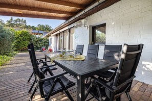 Une terrasse extérieure avec une table et des chaises en bois, un sol en briques et un mur en briques blanches.