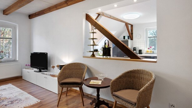Una sala de estar moderna con televisor, sillas de mimbre, una mesita y una cocina visible a través de una ventana.