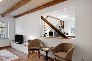 A modern living room with a television, wicker chairs, a small table, and a kitchen visible through a window.