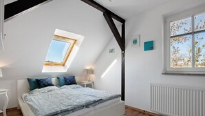 Un dormitorio con techo inclinado, un tragaluz, una cama con ropa de cama estampada, una mesita de noche, una ventana con vistas y un radiador.