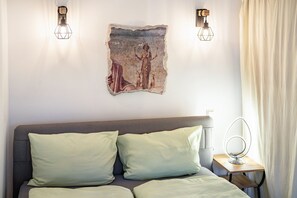 Ein Schlafzimmer mit Bett, Kissen, Wandbild, zwei Wandlampen, einem Nachttisch mit Spiegel und einem Vorhang.