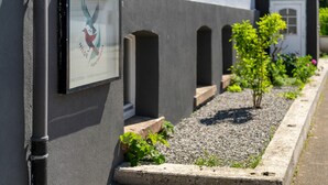 En bygning med en grå facade, et rammet skilt med et fuglelogo, og et lille haveområde med grus og beplantning.