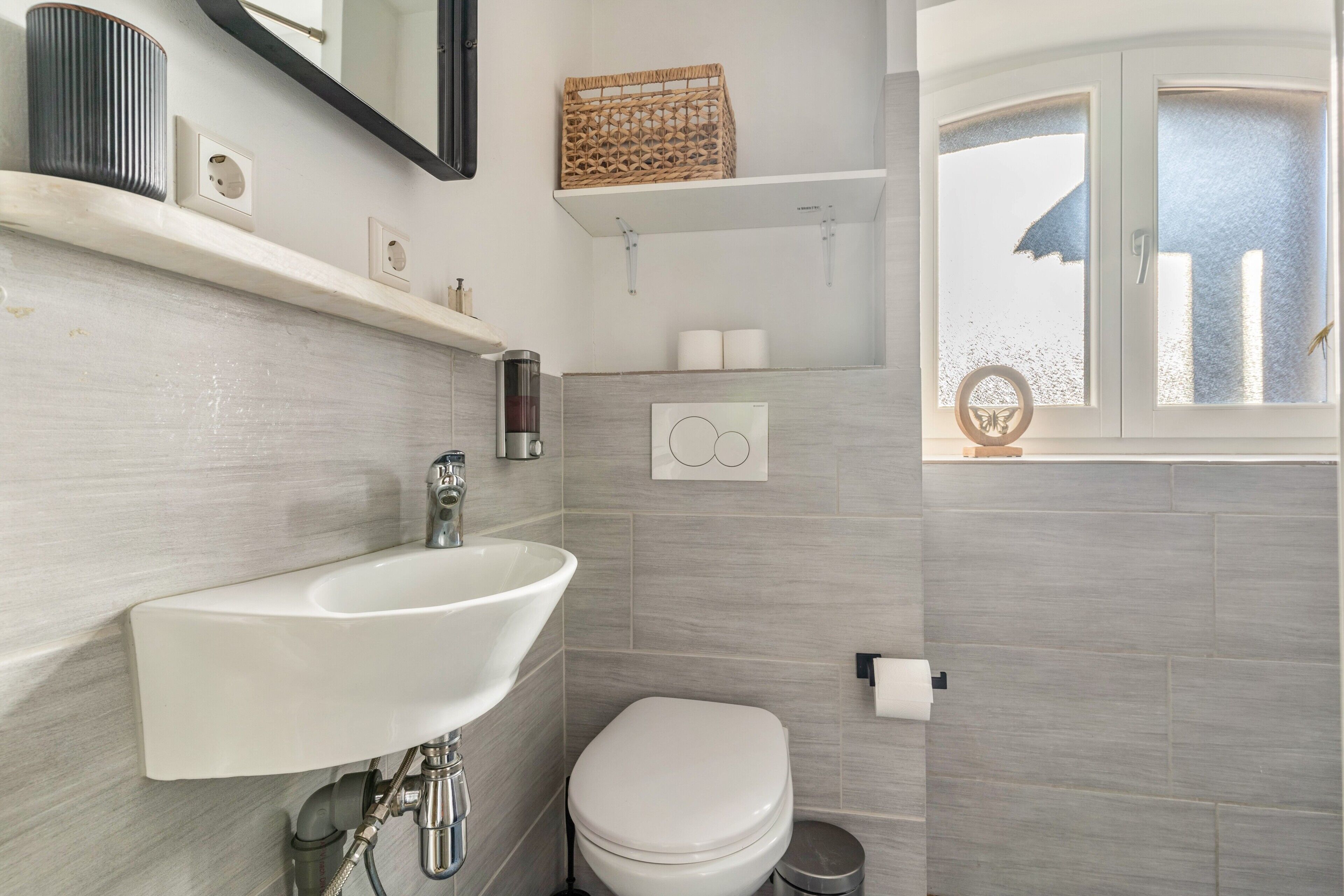A bathroom with a white sink, toilet, and shower area. There are shelves above the sink and a window with frosted glass.