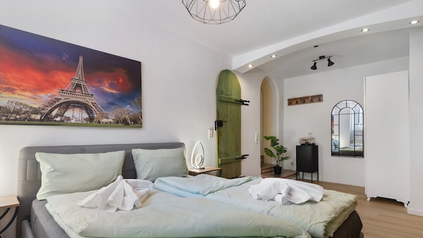 Een slaapkamer met een bed, een schilderij van de Eiffeltoren, een houten deur, een spiegel en een kamerplant.