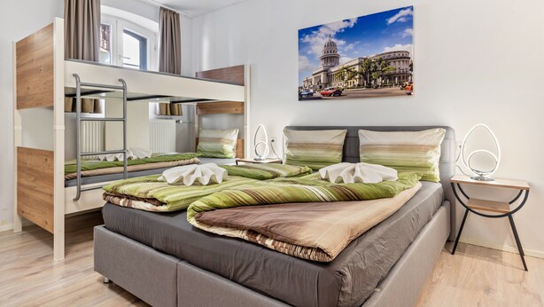 Una cama litera con escritorio y silla, una cama con almohadas, un cuadro en la pared y una ventana con cortinas.