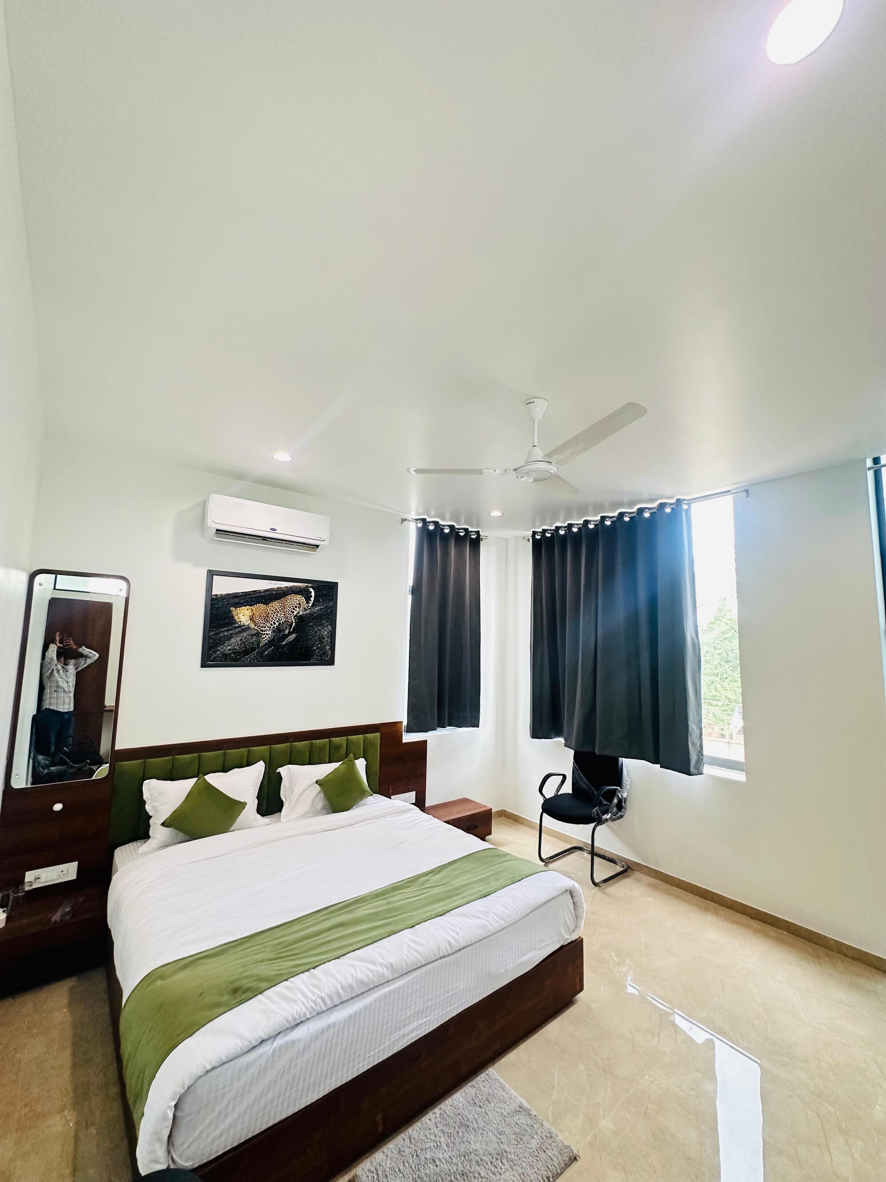 Una habitación de hotel con cama, ventilador de techo, equipo de aire acondicionado, espejo, un cuadro de un leopardo y dos ventanas con cortinas.