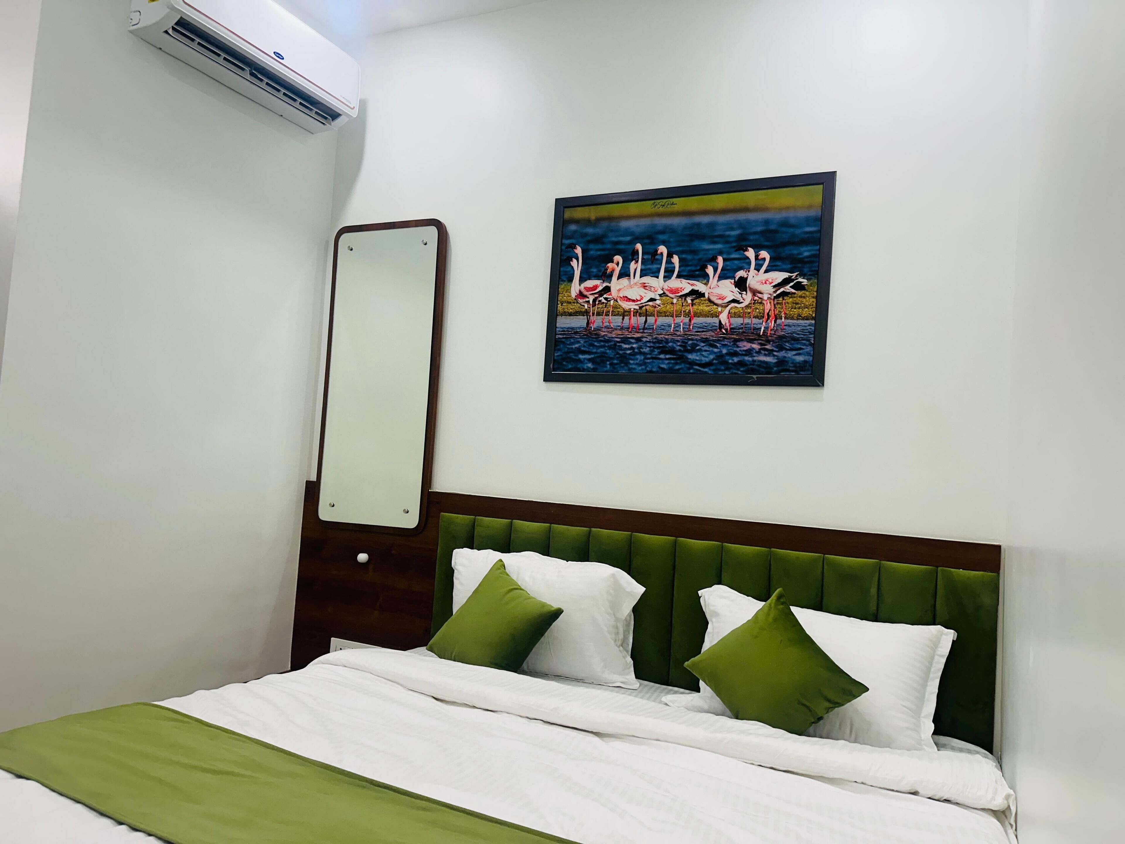 Une chambre d’hôtel avec un lit, une literie verte et blanche, une tête de lit verte, un miroir fixé au mur et un tableau représentant des flamants roses.