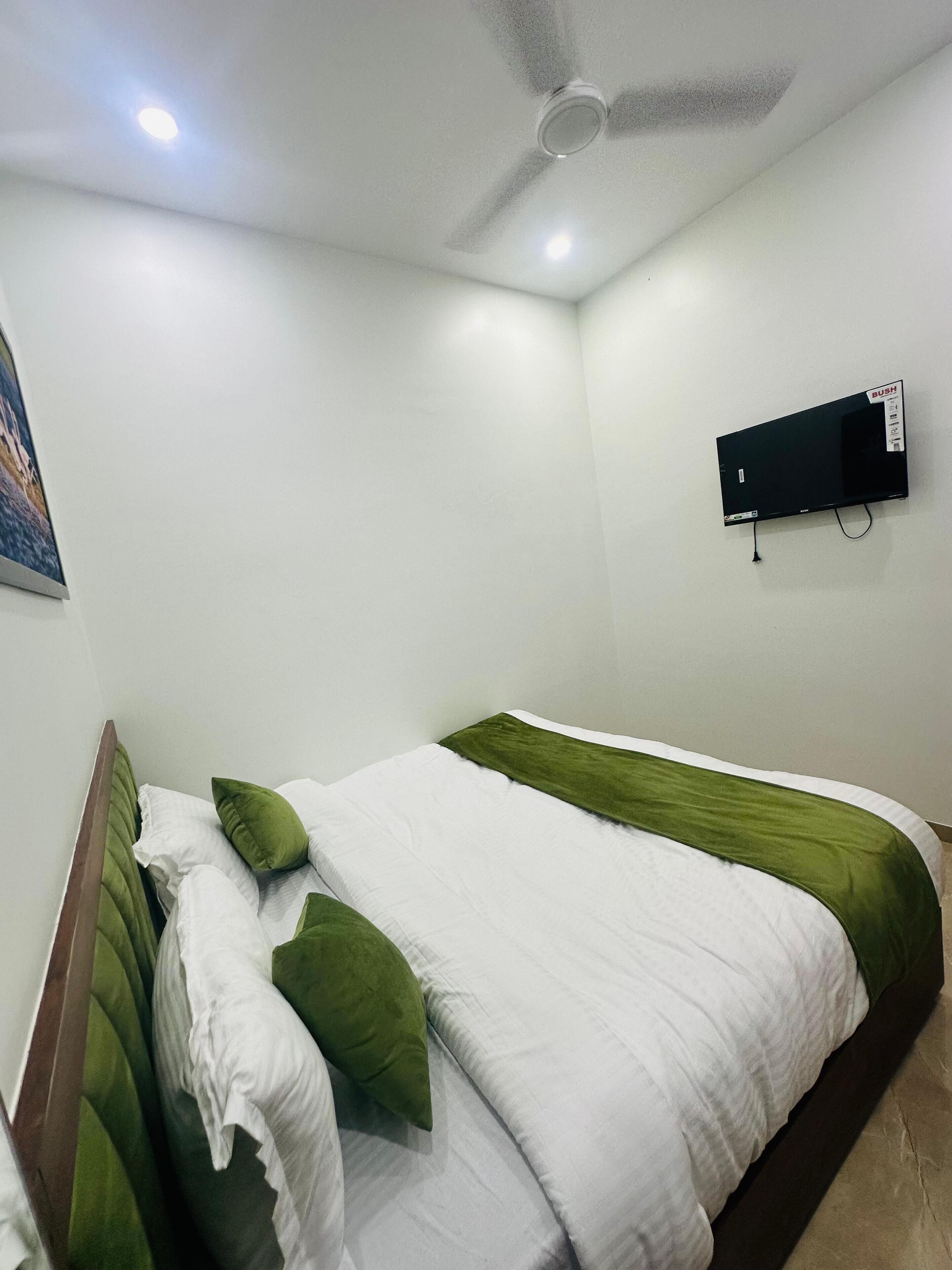 A bedroom with a bed, green and white bedding, a ceiling fan, a mounted TV, and a painting on the wall.