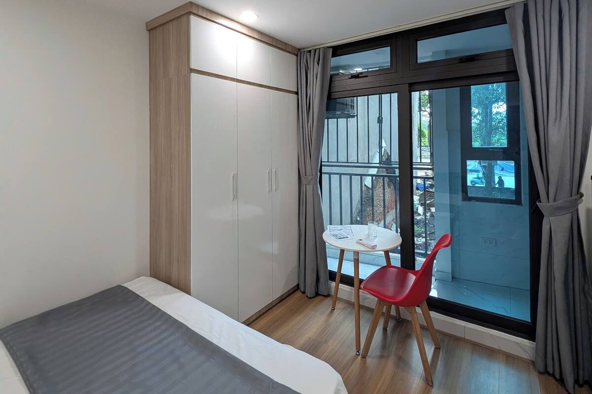 A modern bedroom with a bed, a wardrobe, a small table with a chair, and a balcony with a view.