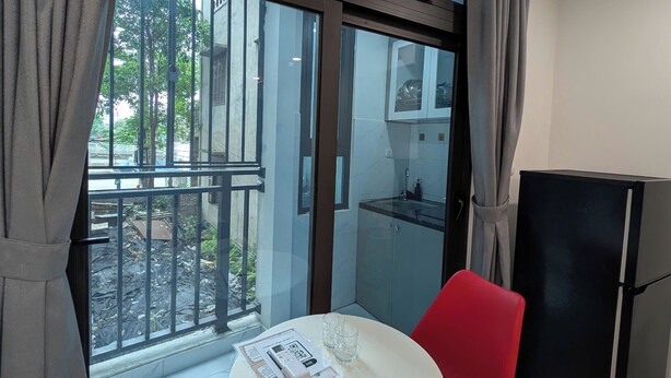 Une petite pièce avec un balcon, une table blanche et une chaise rouge, un réfrigérateur noir et une fenêtre avec des rideaux.