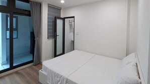 Une chambre avec un lit simple, un plancher en bois, une porte-fenêtre coulissante donnant sur un balcon et une petite table de chevet.