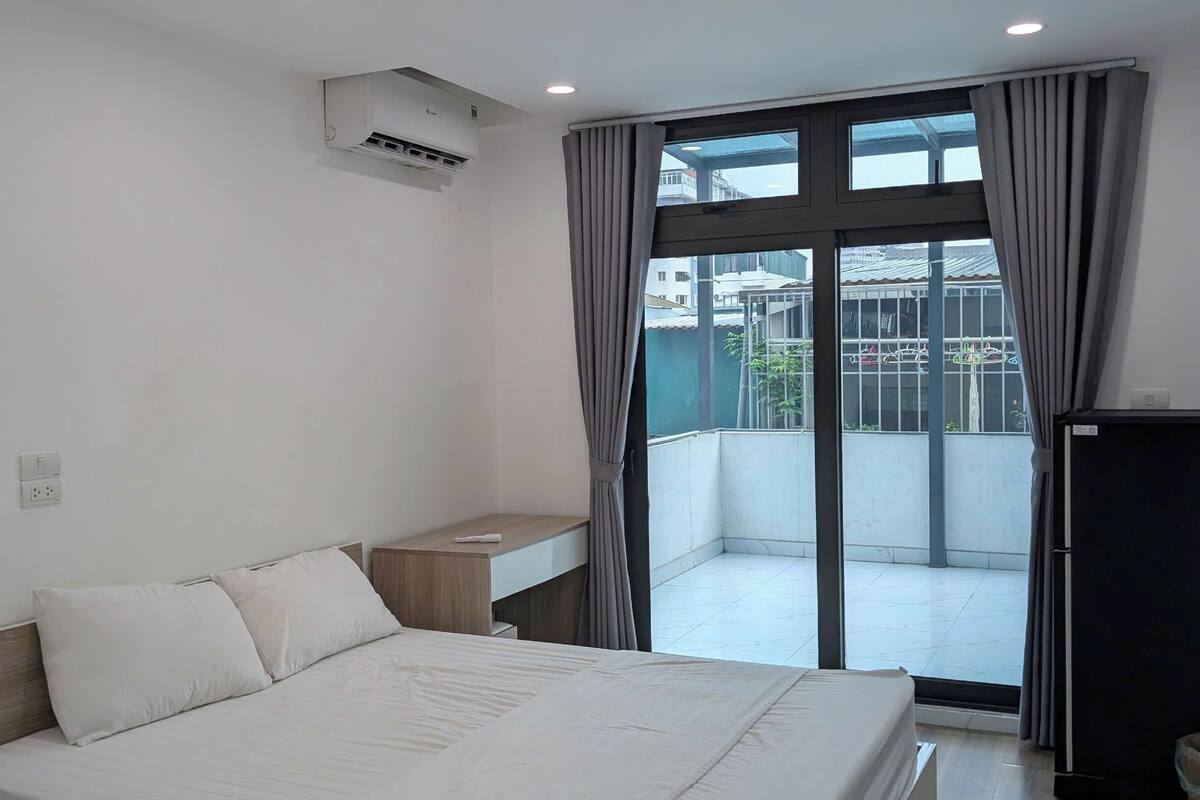 A modern bedroom with a large bed, a small bedside table, an air conditioning unit, and a view of a balcony.