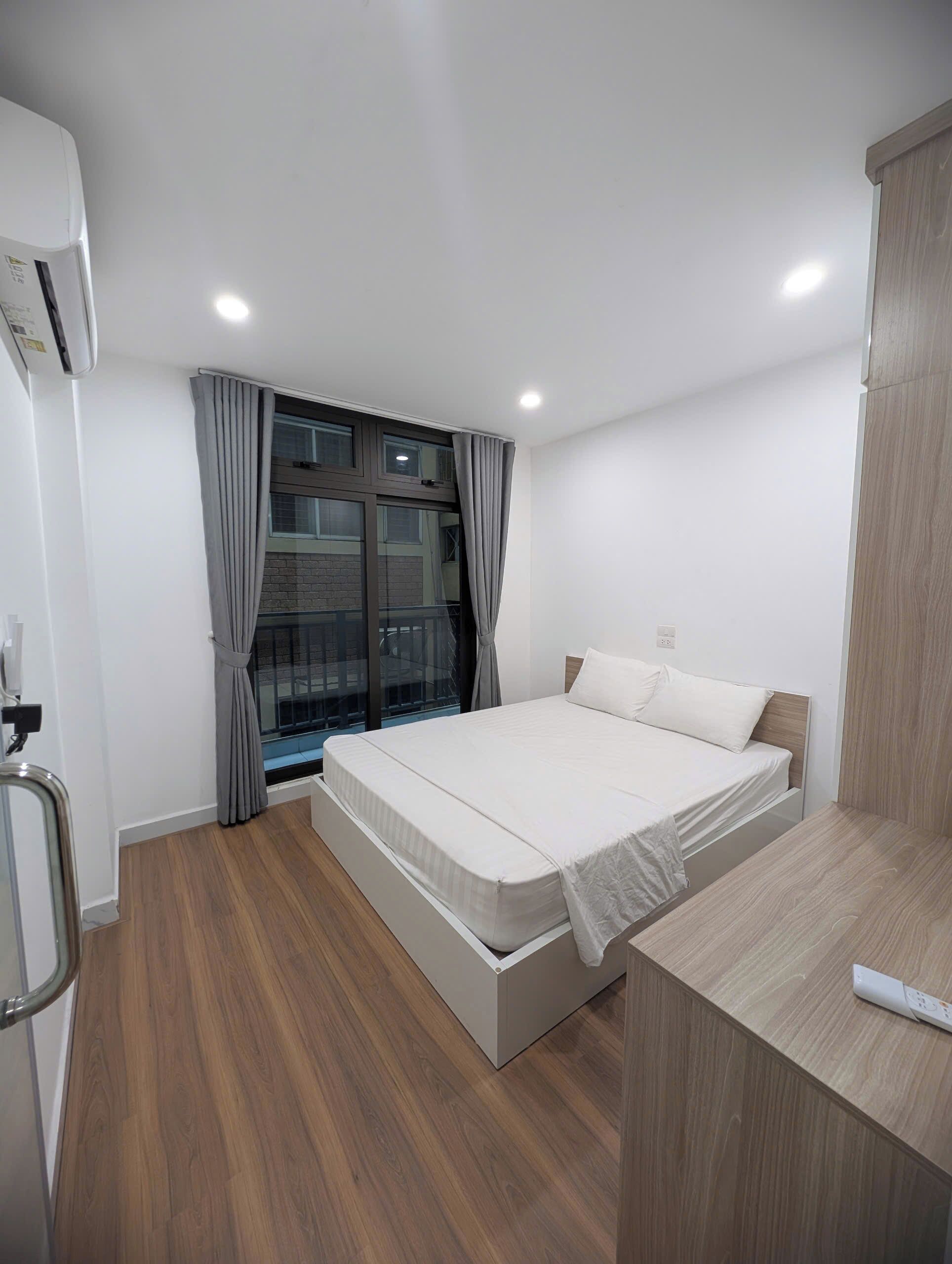 A modern bedroom with a bed, a window with curtains, a wooden floor, and a built-in desk with a remote control.