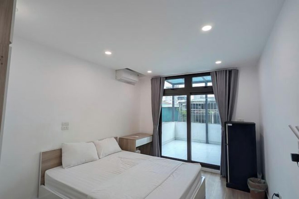 A modern bedroom with a large bed, a small bedside table, a built-in air conditioning unit, a sliding glass door to a balcony, and a black refrigerator.