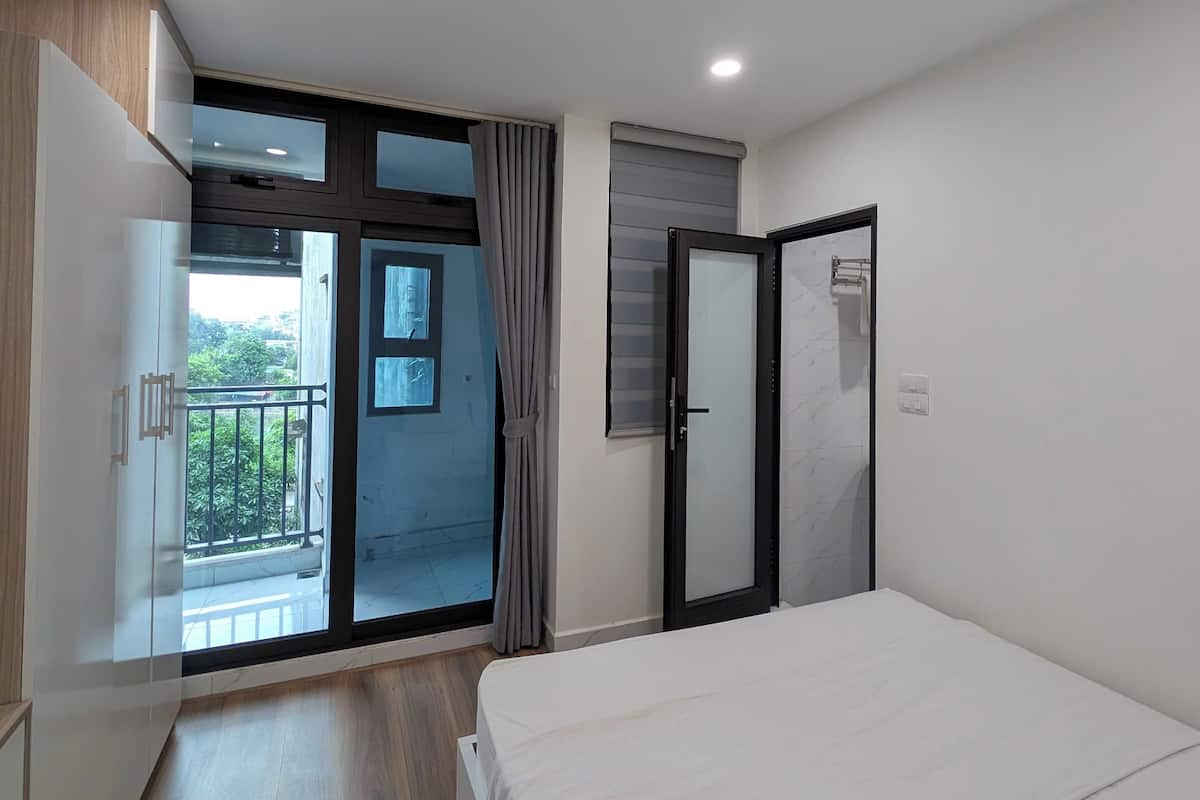 A modern bedroom with a large bed, a sliding glass door to a balcony, and a view of greenery outside.