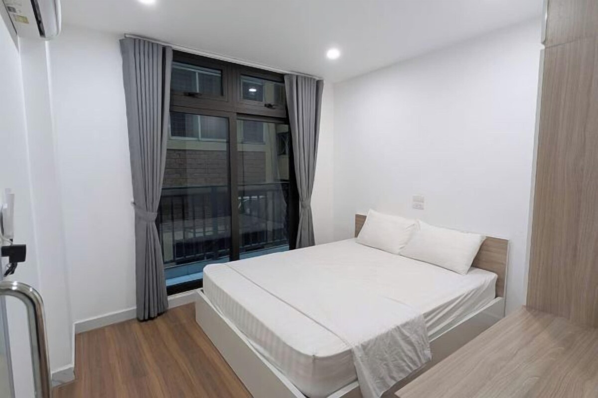 A bedroom with a bed, a window with curtains, a wooden floor, and a wall-mounted air conditioning unit.