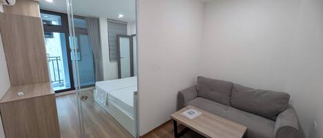 A modern room with a sliding glass door leading to a balcony, a grey sofa, a wooden coffee table, and a small wooden shelf.