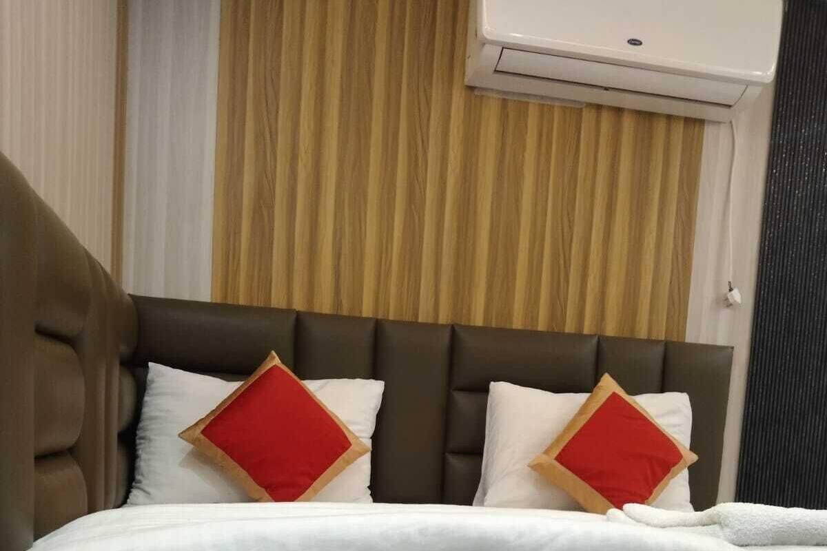 Un lit bien fait, avec une literie blanche et deux coussins rouges et beige, une tête de lit marron recouverte de tissu et un climatiseur fixé au mur.