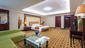 Quarto de hotel com cama, sofá verde, mesa de centro de vidro, escrivaninha com luminária e um espelho grande.