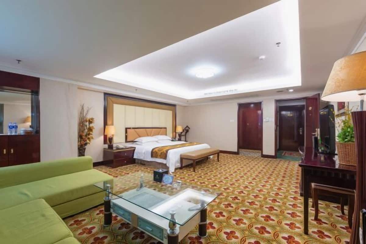 Une chambre d’hôtel avec un lit, un canapé vert, une table basse en verre, un bureau avec une lampe et un grand miroir.