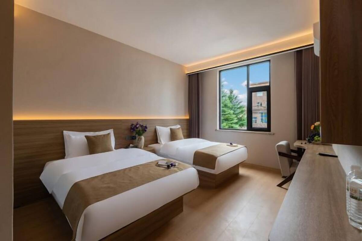 A hotel room with two beds, a desk, and a window with a view of greenery.