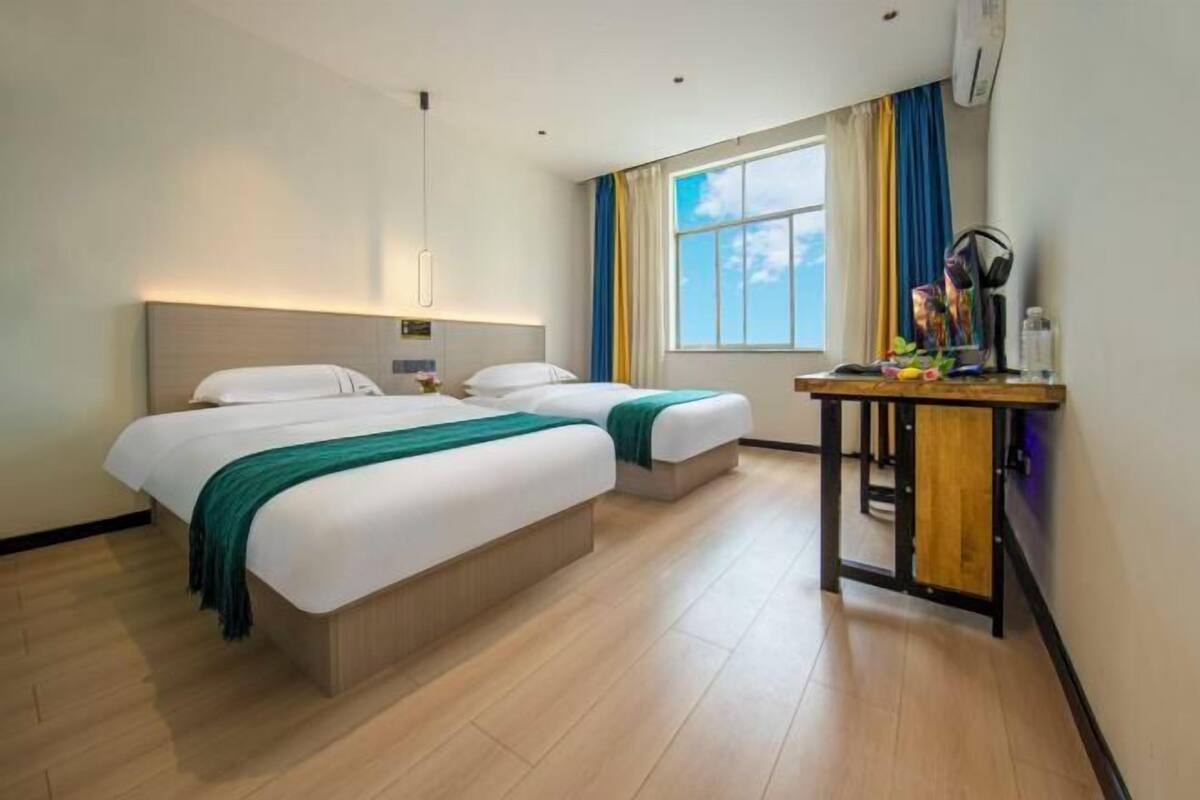 A hotel room with two beds, a desk with a chair, a window with curtains, and an air conditioning unit.