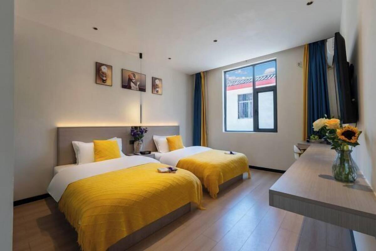 A hotel room with two beds, a desk, a window with curtains, and artwork on the wall.