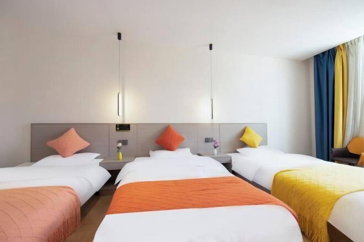 A hotel room with three beds, a nightstand with a lamp, and a view of the city.