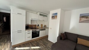 Et kompakt køkken med hvide skabe, en indbygget ovn og en mikrobølgeovn. En træbordplade med et knivdisplay og en lille vask. En mørk sofa er i det tilstødende opholdsområde.