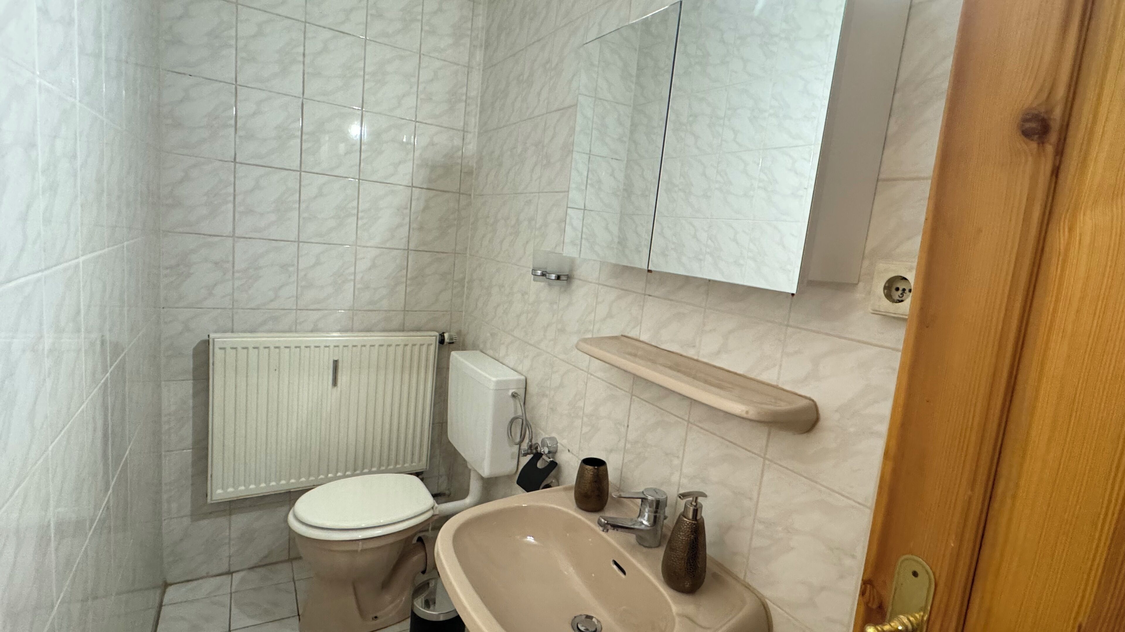 A bathroom with a toilet, sink, mirror, and a wooden shelf.
