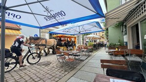 En brostensbelagt gade med udendørs siddepladser under blå parasoller, en hestevogn og en cyklist.