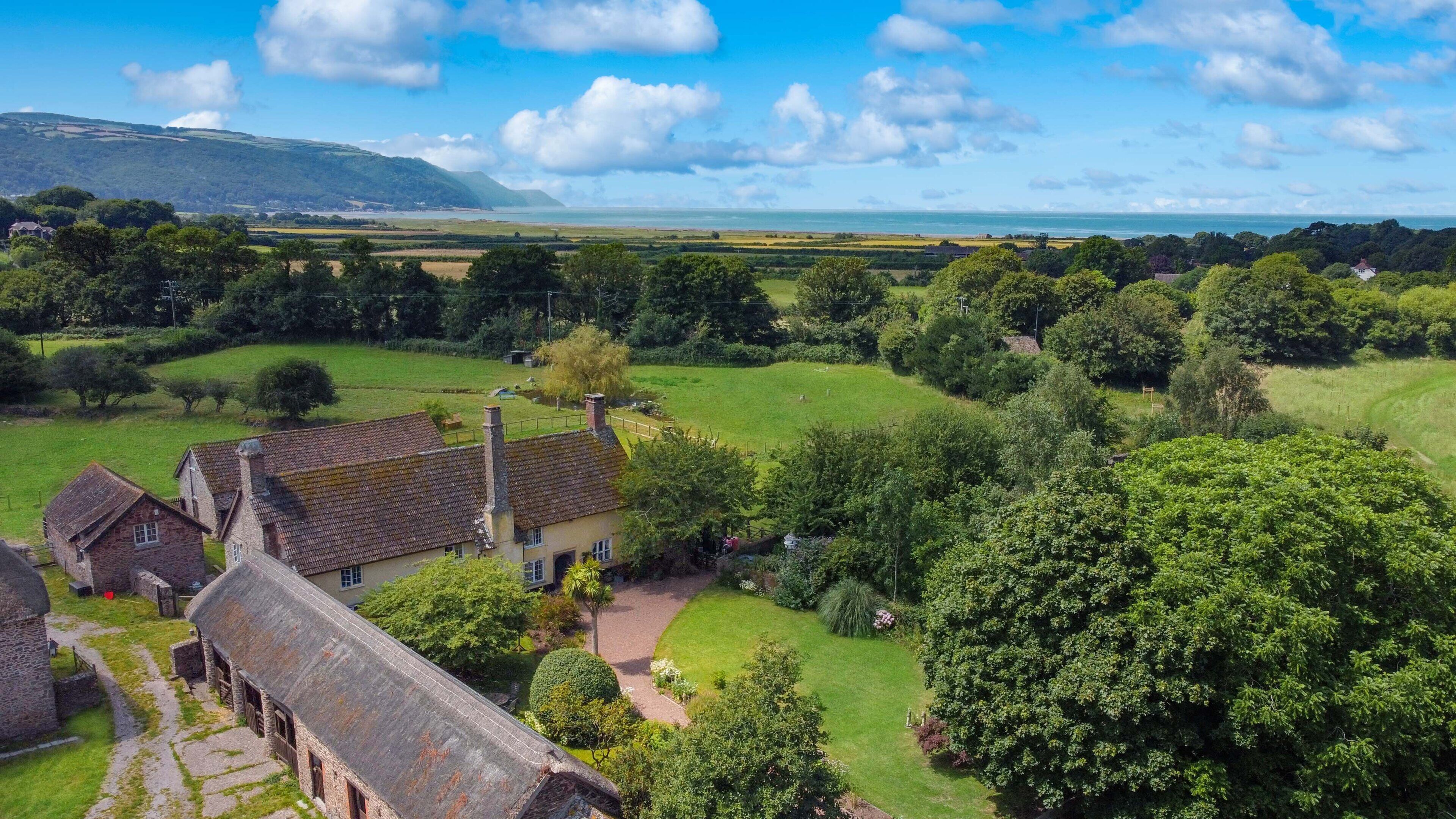 A rural landscape with traditional stone buildings, a large grassy field, and a distant view of the sea.