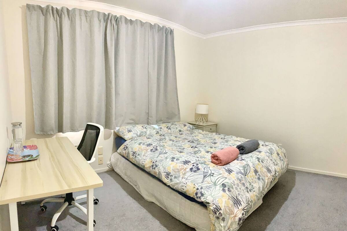 Um quarto pequeno e bem organizado, com uma cama de solteiro, uma escrivaninha e uma cadeira.