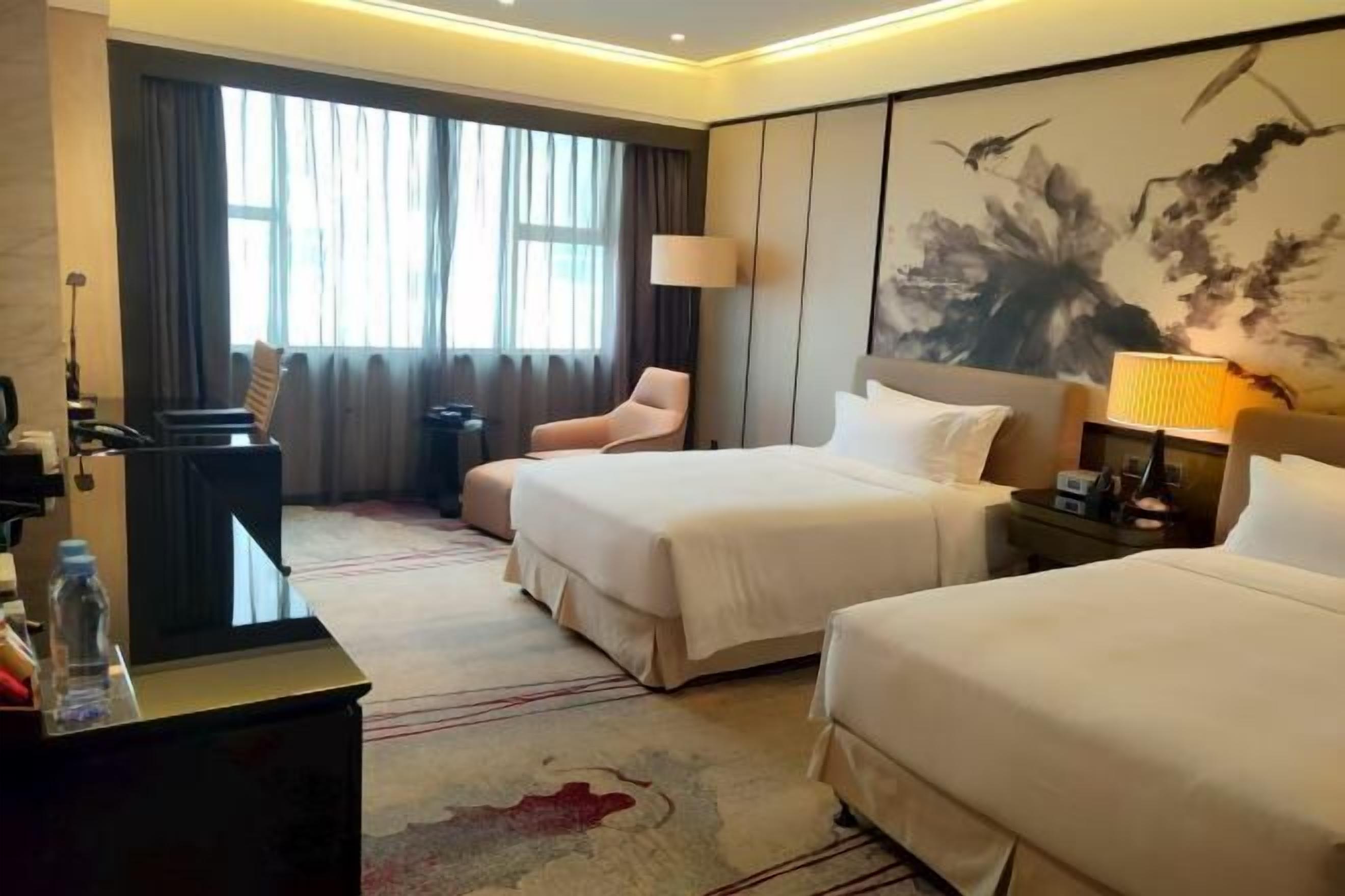 Habitación de hotel con dos camas, un escritorio con lámpara, una silla y un ventanal con cortinas.