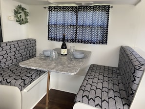 Une petite cuisine équipée comprenant une table, deux chaises, une bouteille de vin et une fenêtre agrémentée de rideaux à motifs.