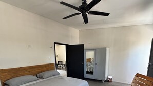 Une chambre à coucher comprenant un lit, une chaise en bois, un ventilateur de plafond et une porte donnant sur une autre pièce.