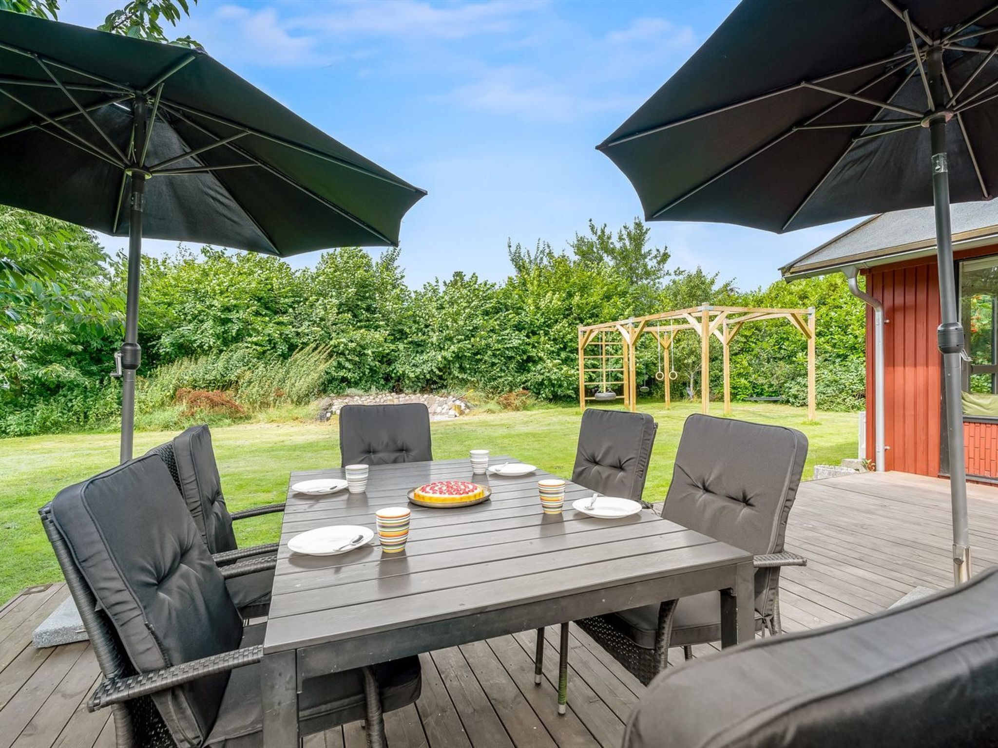 An outdoor dining area with a wooden deck, a table set for four, and two large umbrellas providing shade.
