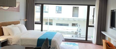 A modern hotel room with a large bed, a desk, a chair, and a TV. There is a balcony with a view of the city.