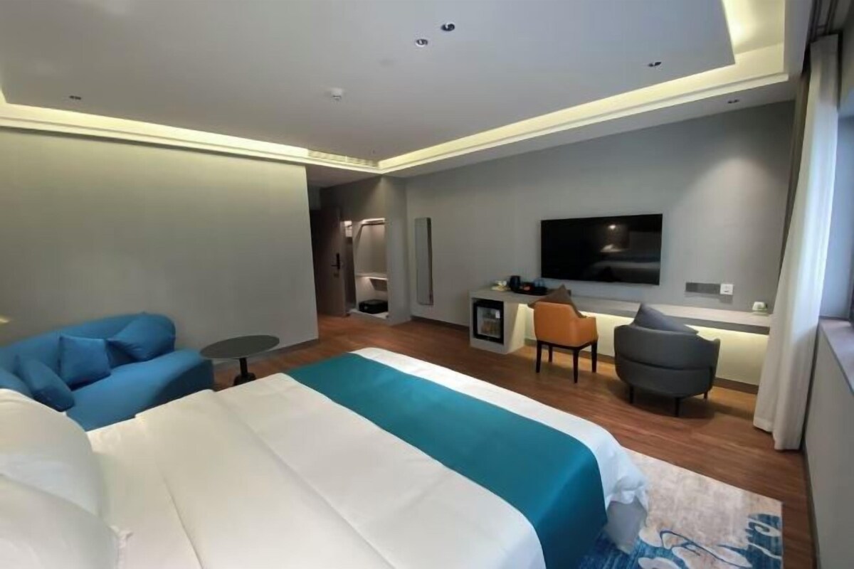 A modern hotel room with a large bed, a seating area with a sofa and armchairs, a small desk, and a TV mounted on the wall.