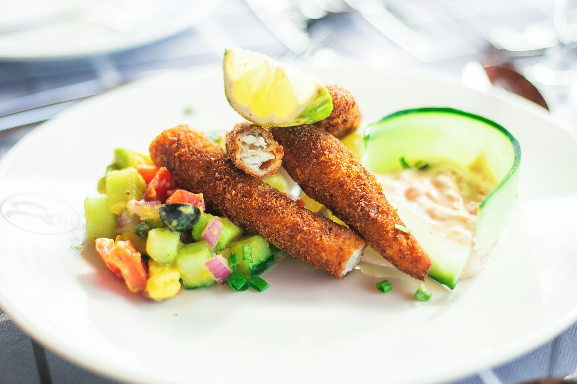 En tallerken med paneret fiskefilet, en agurkesalat og en skive agurk.