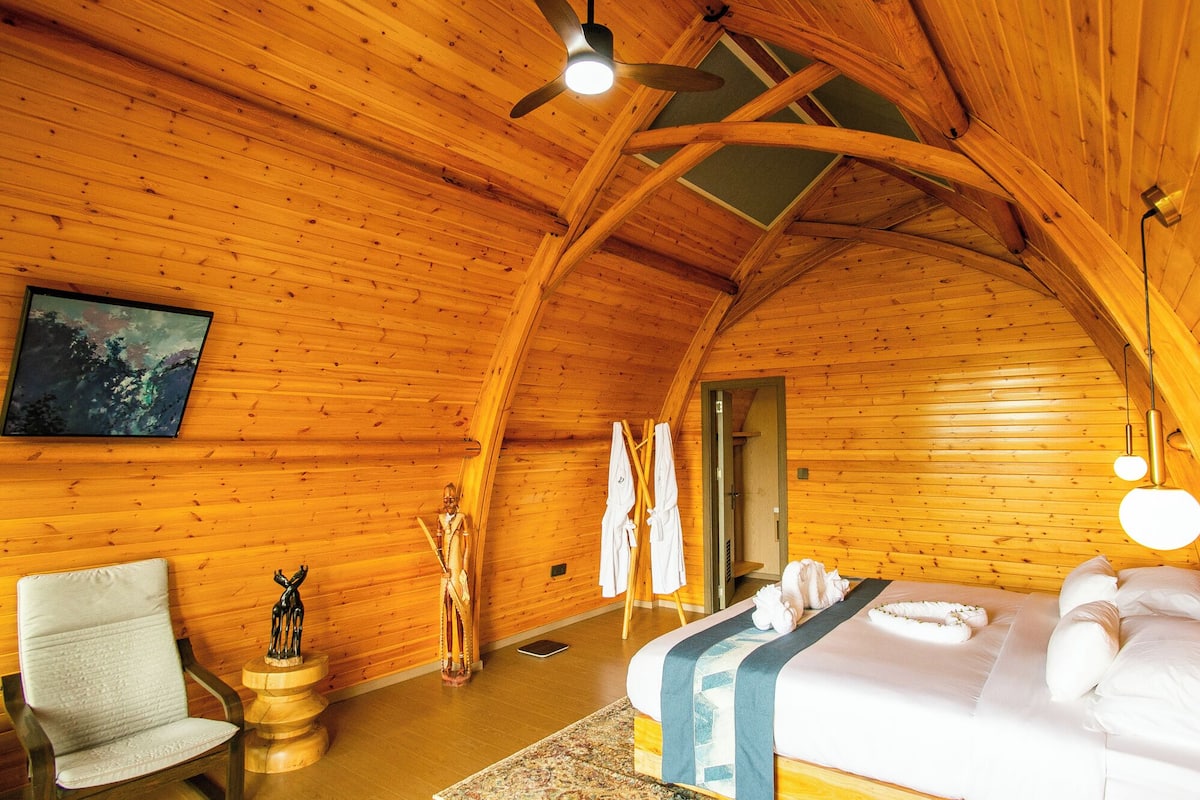 A cozy wooden cabin room with a bed, a chair, a painting, and a ceiling fan.