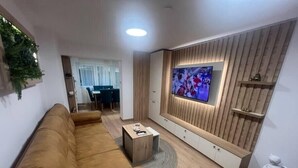 Moderni olohuone, jossa on ruskea sohva, sohvapöytä, puiseen seinään asennettu televisio ja taustalla ruokailutila.