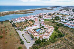 Vista aérea de una zona residencial costera con edificios blancos, una piscina central y una playa al fondo.