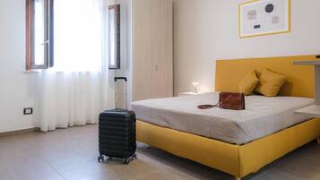 Una habitación con cama, maleta, ventana con cortinas, un cuadro en la pared y una mesita de noche con lámpara.